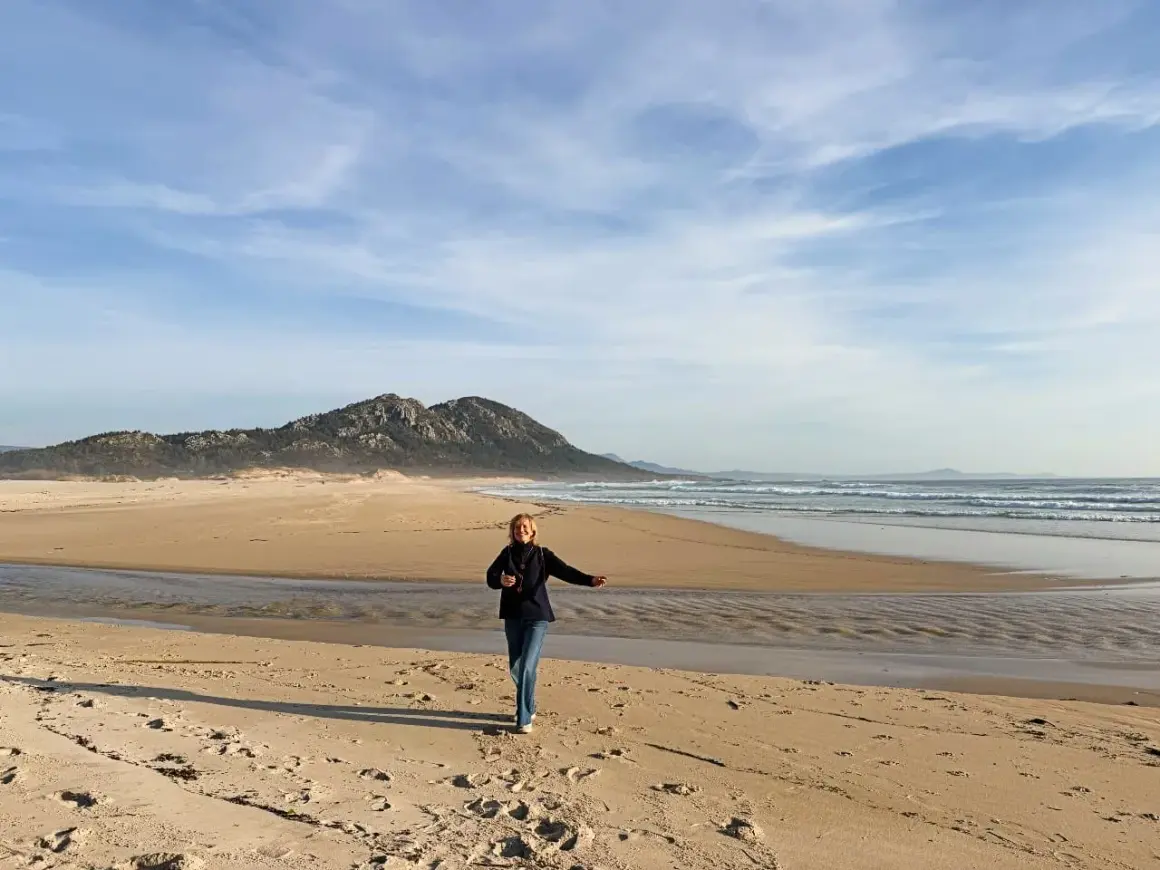 Mujer corre en la playa de Muros, Galicia, con montañas al fondo y el mar azul.