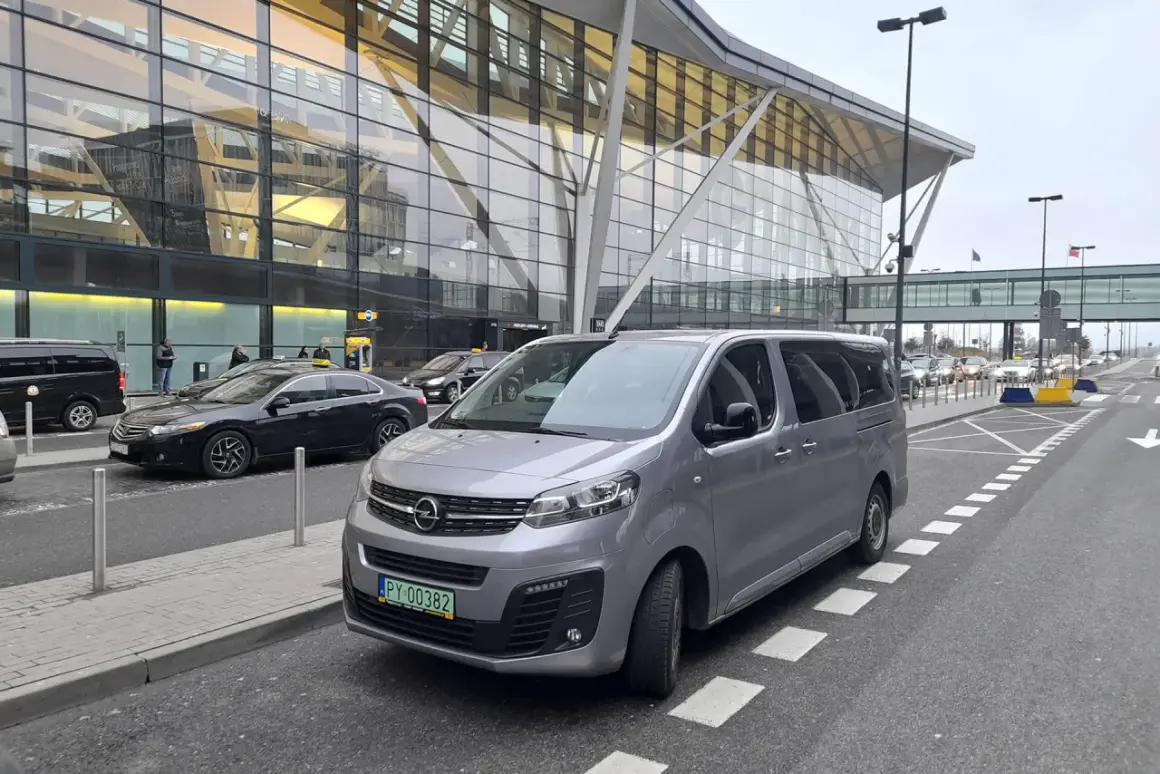 Szary van czeka na pasażerów przed nowoczesnym terminalem lotniska. Może to być transport z Koszalina do Gdańska.