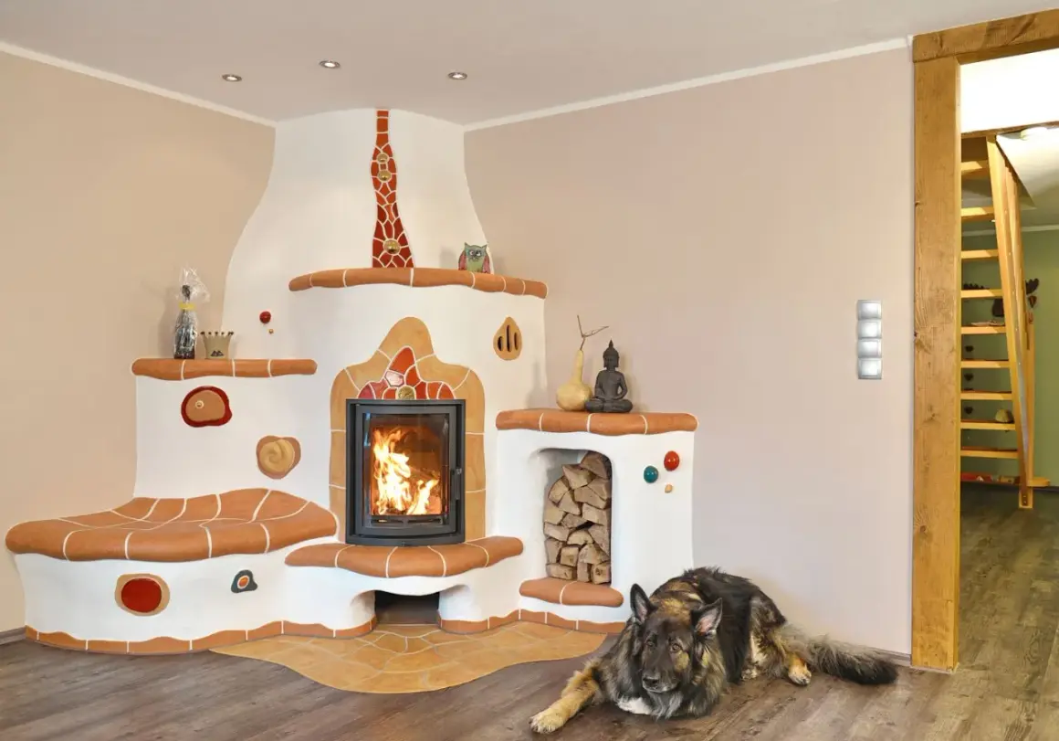 Ein Hund liegt vor einem großen, weißen Kachelofen mit integriertem Kamin. Der Ofen hat eine Sitzbank und ist mit bunten Kacheln verziert. Der Unterschied zwischen Kamin und Ofen wird hier durch die massive Bauweise und die Heizfunktion des Ofens deutl...