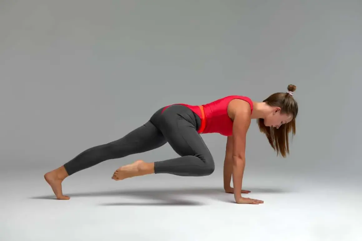 Kobieta w czerwonej bluzce i szarych legginsach wykonuje dynamiczny stretching, przyciągając kolano do klatki piersiowej w pozycji deski.