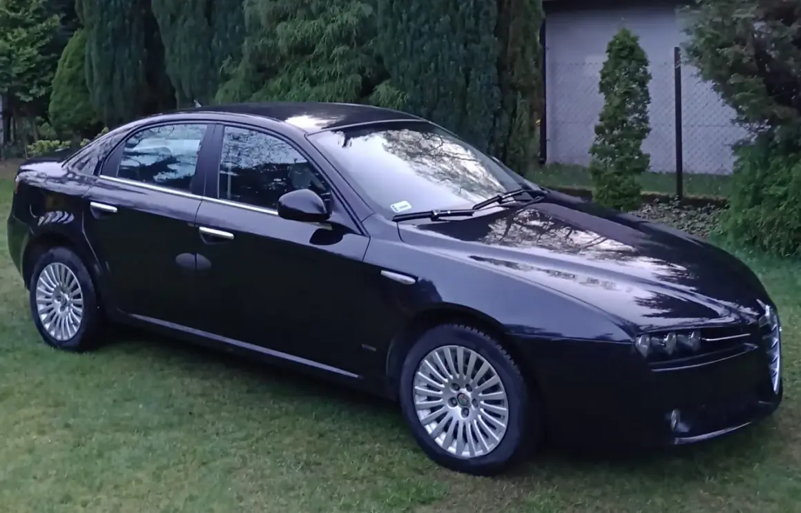 Elegancka, czarna Alfa Romeo 159 stoi na trawniku, otoczona zielenią.