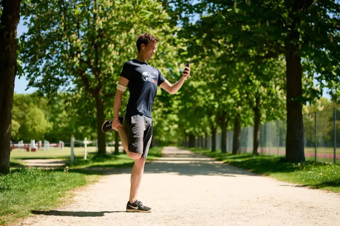 Un homme fait un étirement quadriceps sur un chemin en terre, regardant son téléphone.