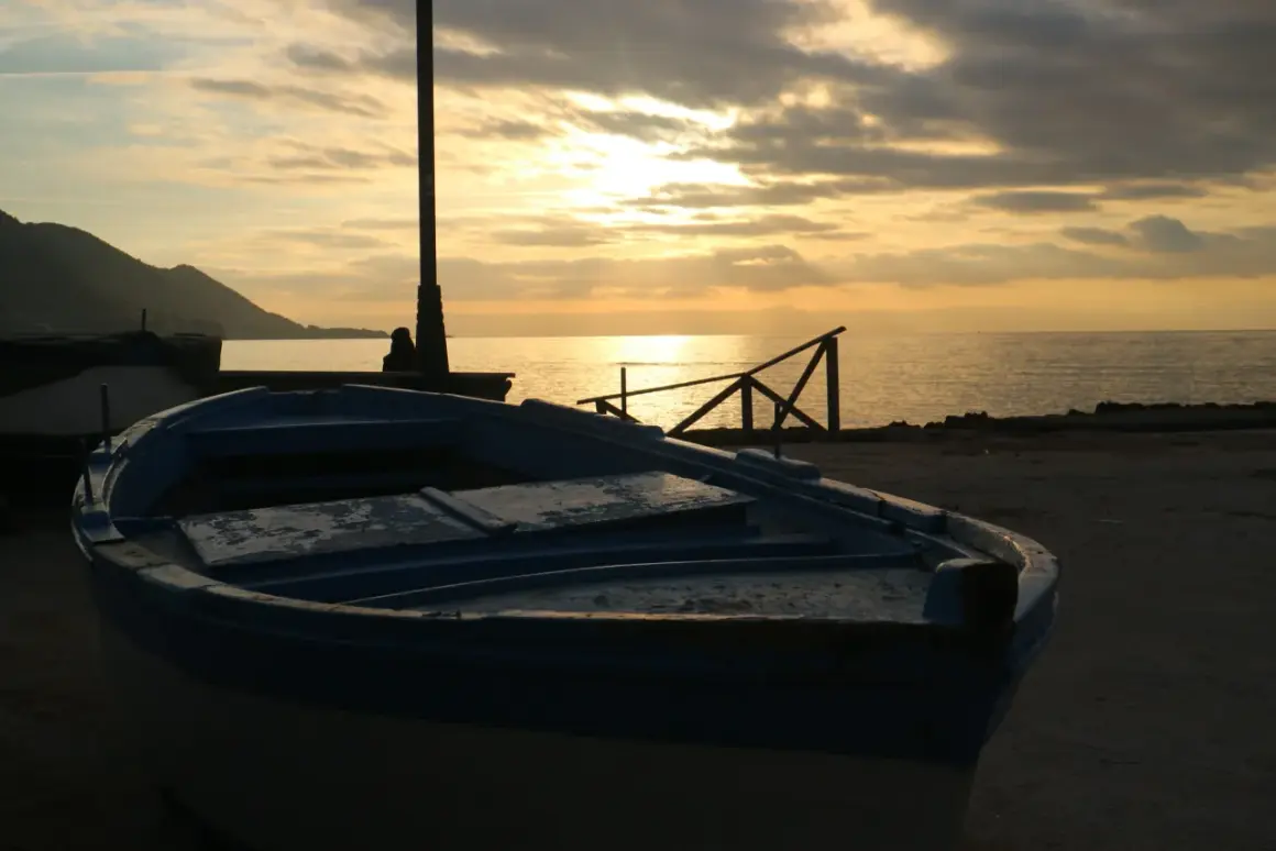 Un'imbarcazione blu riposa sulla riva al tramonto, con il sole che filtra tra le nuvole e illumina il mare calmo.