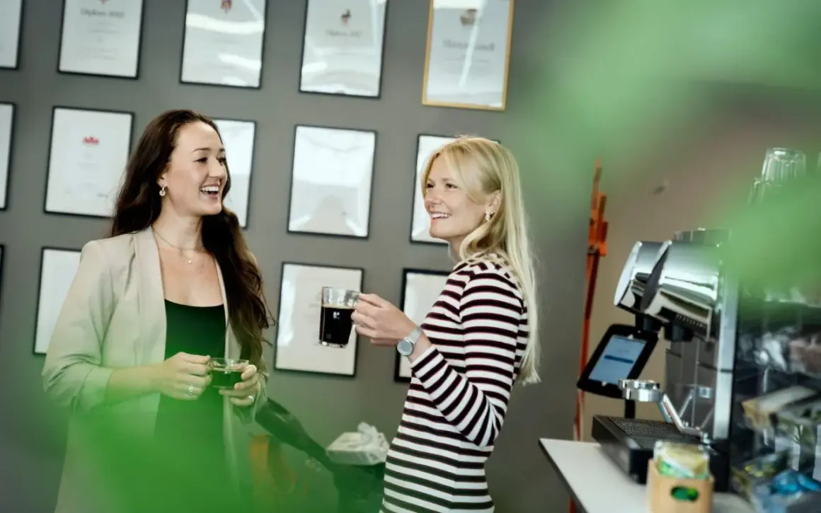 Två kvinnor, en grafisk formgivare, pratar vid en kaffemaskin. Väggen bakom dem är prydd med diplom.