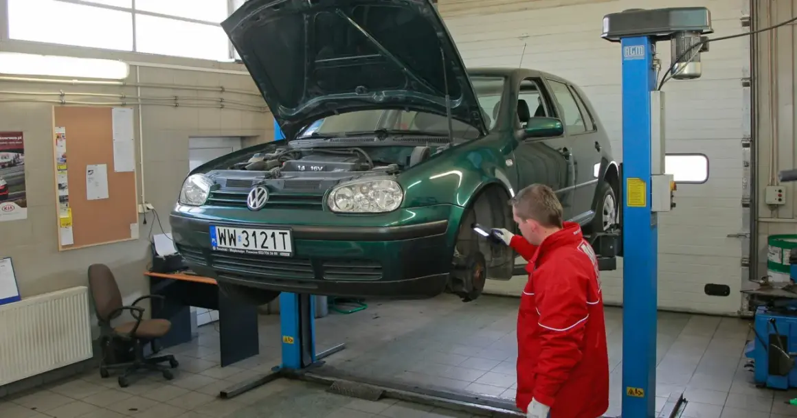 Mechanik w czerwonej kurtce sprawdza podniesiony na podnośniku zielony samochód. To kluczowy moment, by dowiedzieć się, jak dbać o samochód.