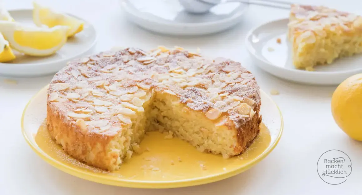 Saftiger Kuchen ohne Mehl, bestreut mit Mandelblättchen und Puderzucker. Dazu Zitronenspalten und eine ganze Zitrone.