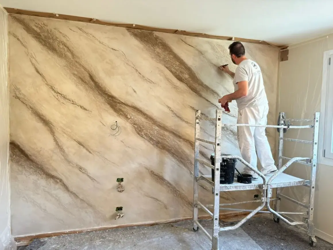 Pittore applica stucco effetto marmo su una parete, creando venature naturali.