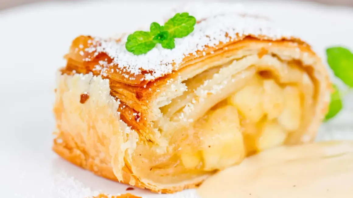 Ein Stück Apfelstrudel mit Puderzucker und Minze, serviert mit Vanillesauce. Perfekt für ein Apfelstrudel Rezept!
