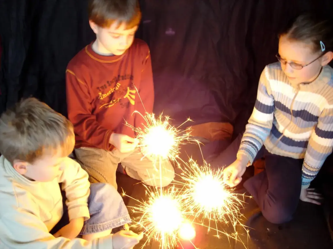 Kinder halten Wunderkerzen. Ein Junge im roten Pullover, ein Mädchen mit Brille und ein kleiner Junge im hellen Pulli genießen den Zauber. Ein schöner Moment, der an die Grundschule erinnert.