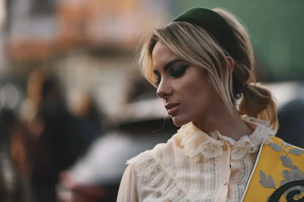 Blonde Frau mit auffälligem Make-up und einem grünen Haarband, das ihre Frisur betont.