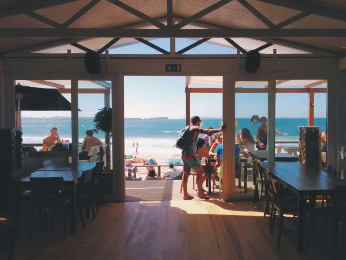 Widok z wnętrza restauracji na plażę i morze. W środku ludzie siedzą przy stołach, na zewnątrz plażowicze odpoczywają. To najlepsza restauracja na Helu.
