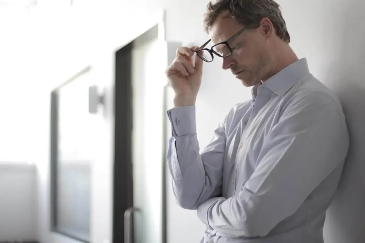 Mann mit Brille, der sich an eine Wand lehnt. Er wirkt nachdenklich, vielleicht kämpft er mit Hitzewallungen.