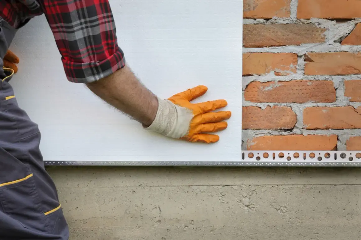 Pose d'isolant blanc sur un mur de briques. La main gantée ajuste la plaque, une étape clé dans la définition de l'isolation.