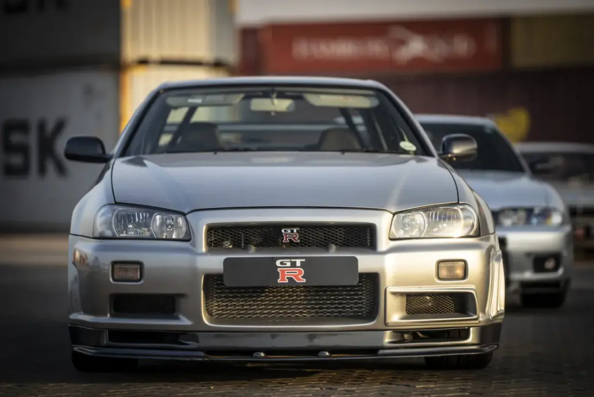 Srebrny Nissan Skyline GT-R R34 z bliska, z innym GT-R w tle. Ikoniczny samochód sportowy.