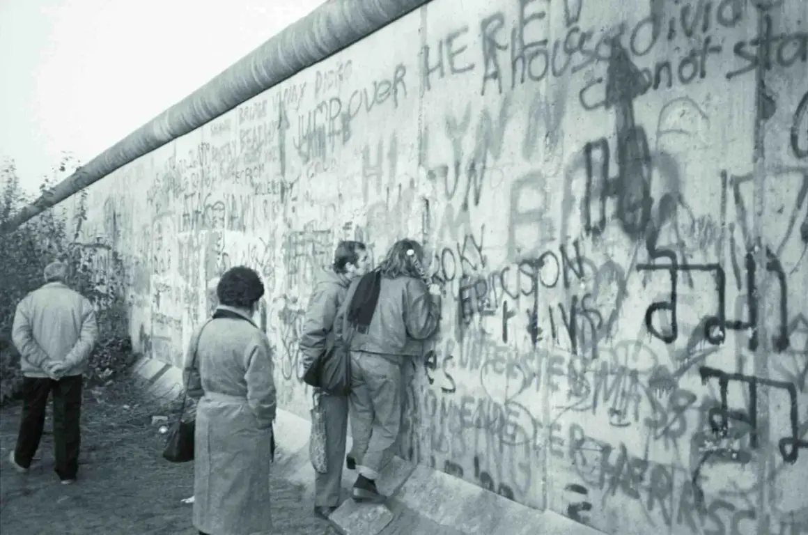Ludzie oglądają graffiti na Murze Berlińskim, gdy zburzono mur. Widać napisy "Jump over here" i "A house divided cannot stand".