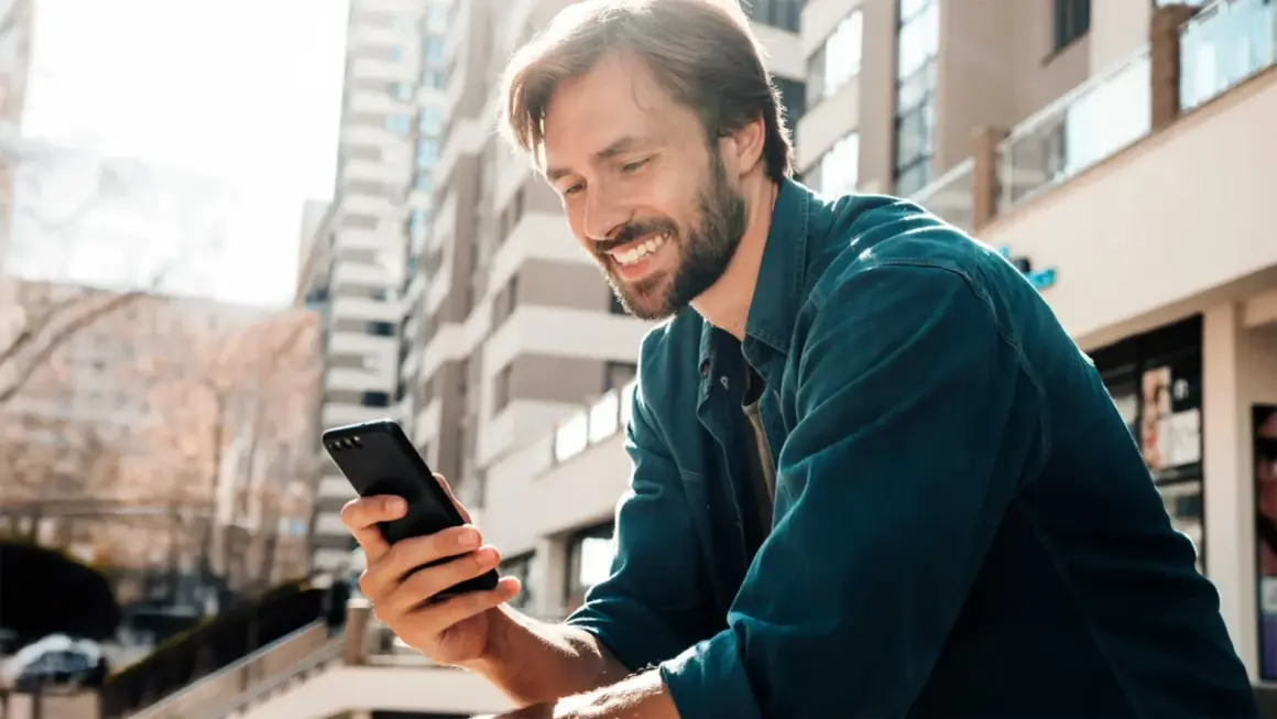 Uśmiechnięty mężczyzna z zarostem trzyma w dłoni czarny telefon, przeglądając coś z zaciekawieniem. W tle nowoczesne budynki.