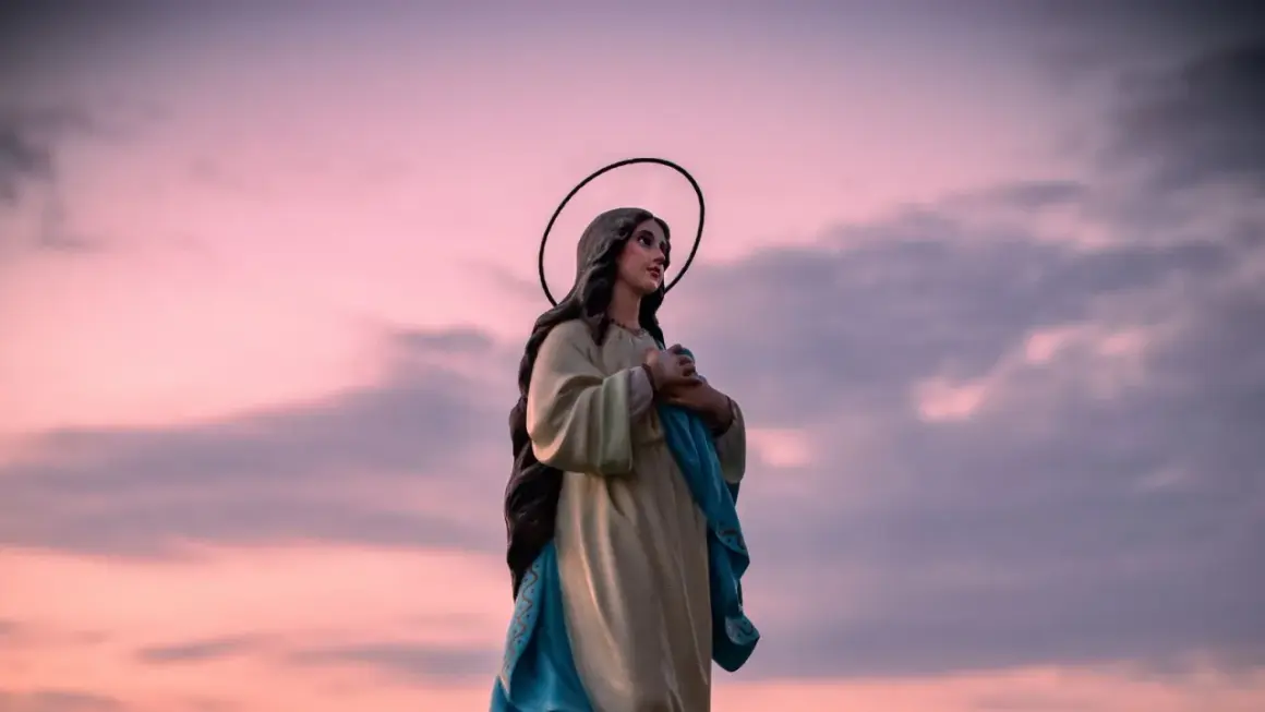 Figurka Matki Boskiej z aureolą na tle różowego nieba. W dłoniach trzyma błękitną szatę, jakby w modlitwie "Anioł Pański".