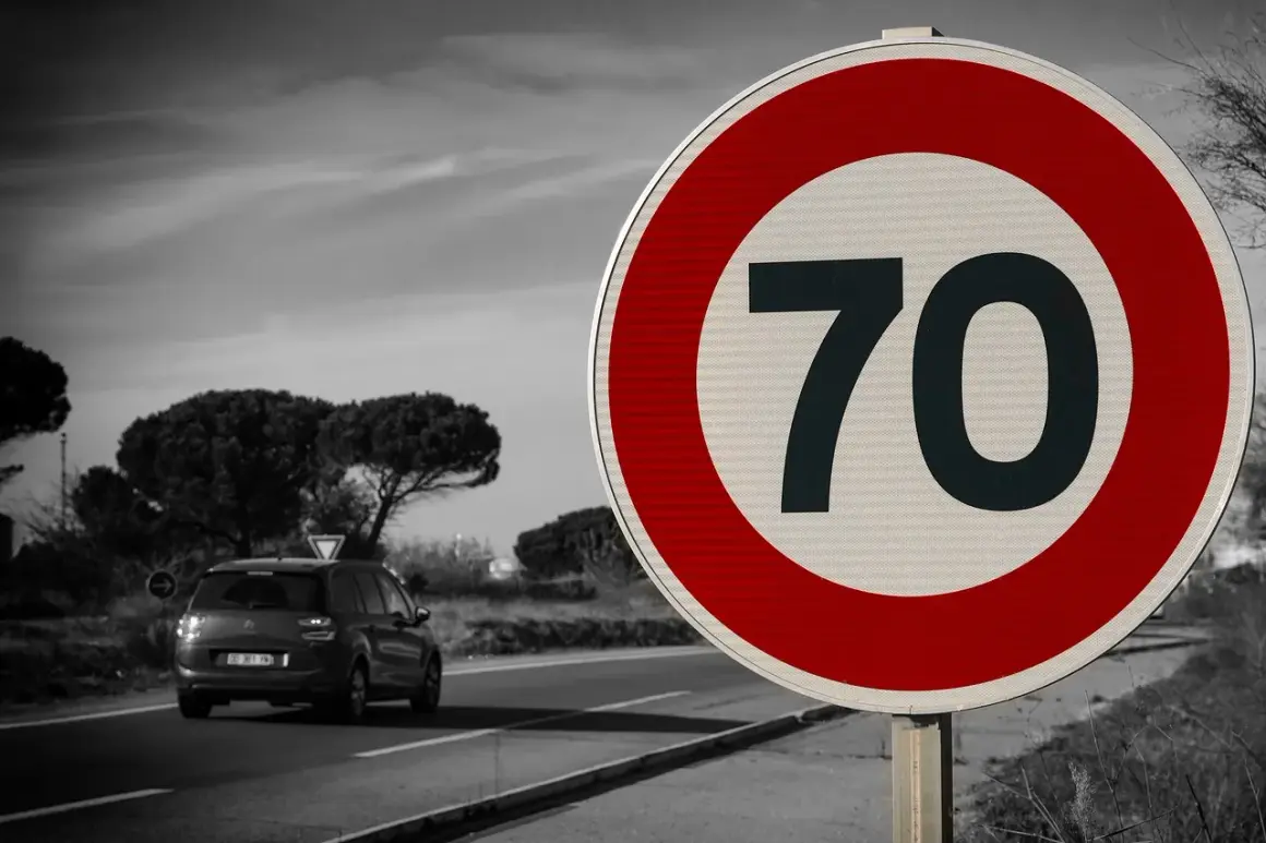 Zbliża się znak ograniczenia prędkości do 70 km/h. Samochód jedzie po drodze w tle.