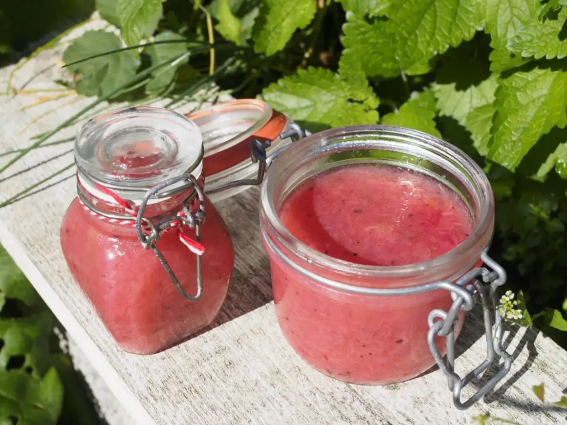 Två glasburkar med rabarbermarmelad med kardemumma står på en träbänk utomhus, omgivna av gröna blad.