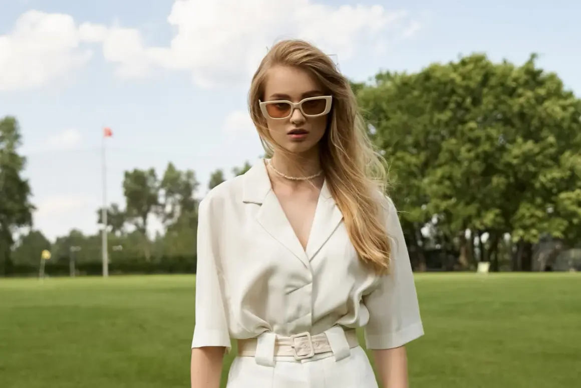 Młoda kobieta w okularach i eleganckim, białym stroju stoi na tle zielonego parku.