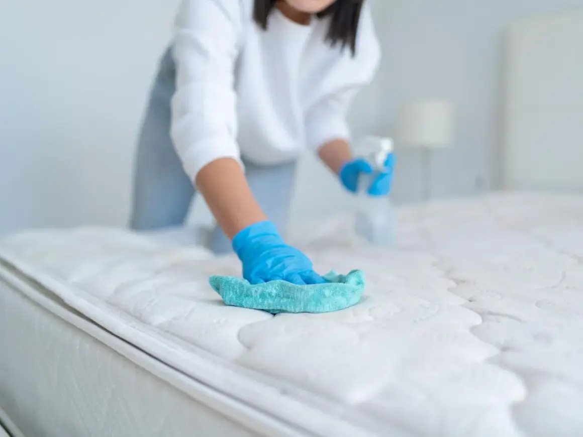 Person mit blauen Handschuhen reinigt die Matratze mit einem Tuch und Sprühflasche.