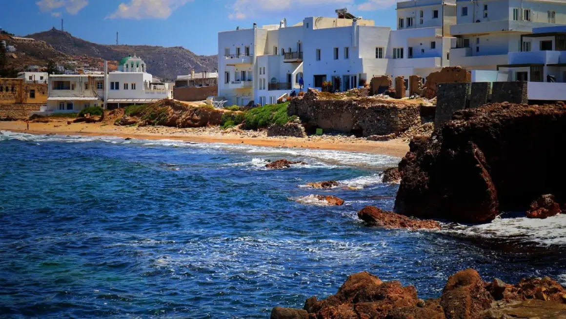 Blågrönt hav sköljer mot klippor och sandstrand. Vita hus med gröna tak reser sig mot en bergig bakgrund på Naxos, Grekland.