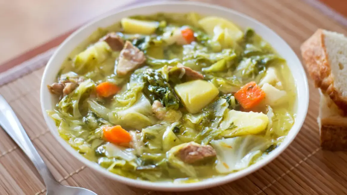 Bol de soupe au chou vert, pommes de terre, carottes et viande, avec du pain à côté.