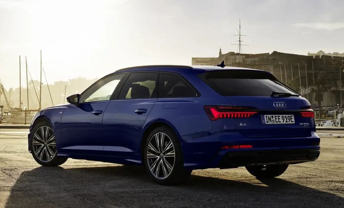 Niebieskie Audi A6 Avant z napisem "50 TFSI e" na klapie bagażnika, zaparkowane przy nabrzeżu z widokiem na zabytkową fortecę i żaglówki.
