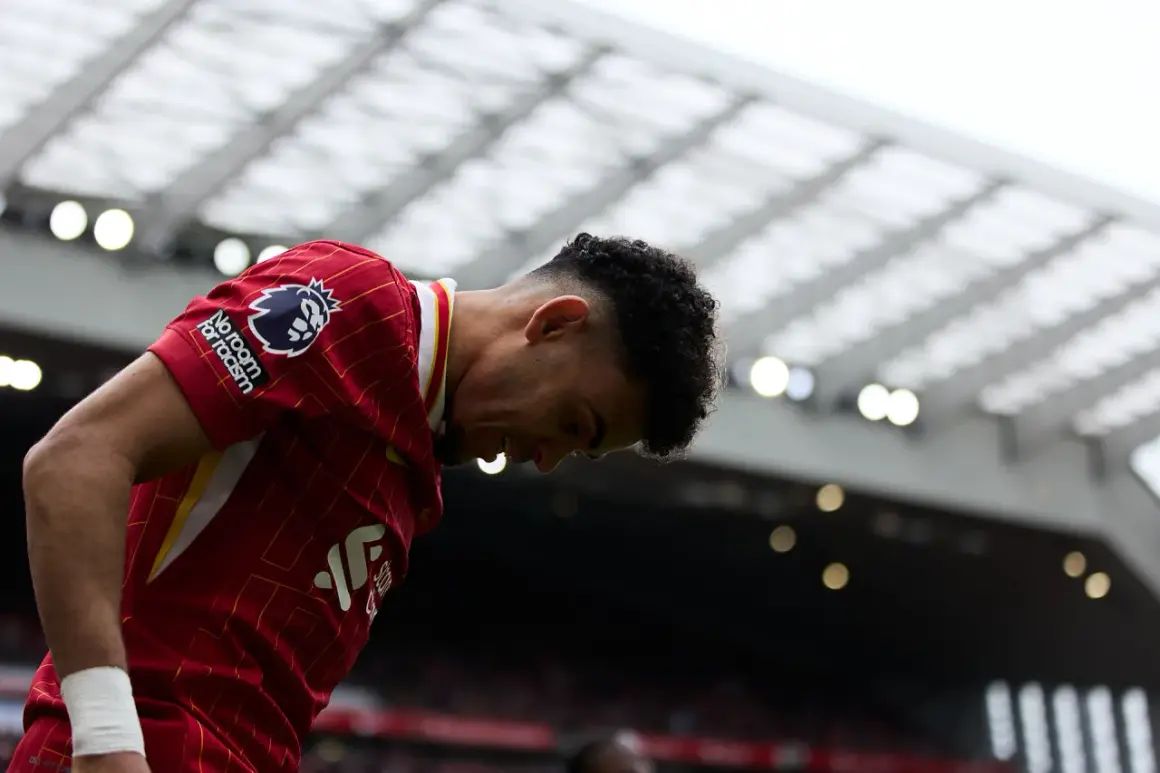 Piłkarz w czerwonej koszulce Liverpoolu z opuszczoną głową, na tle trybun stadionu.