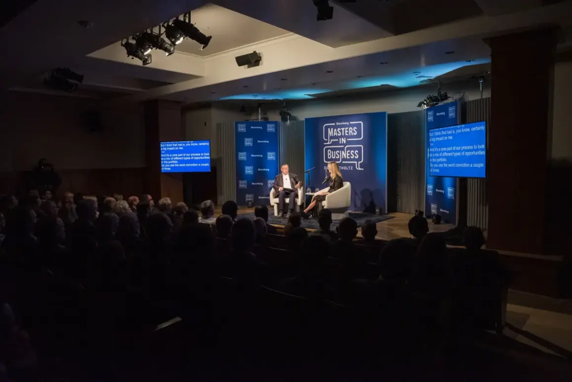 Conference photography captures a panel discussion on a stage with two speakers, audience members in the foreground, and Bloomberg branding.