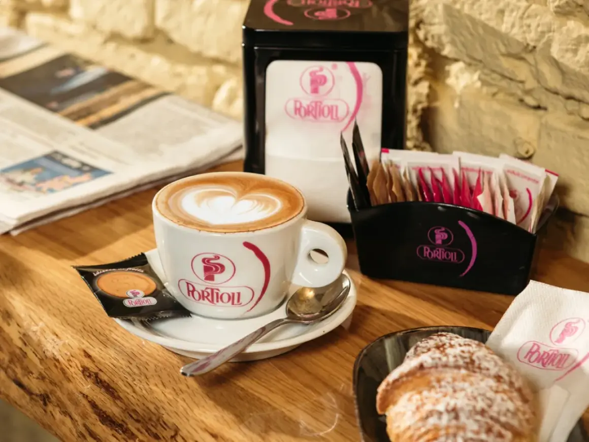 Un cappuccino senza schiuma, con un cuore di latte art, accompagnato da un croissant e un giornale.
