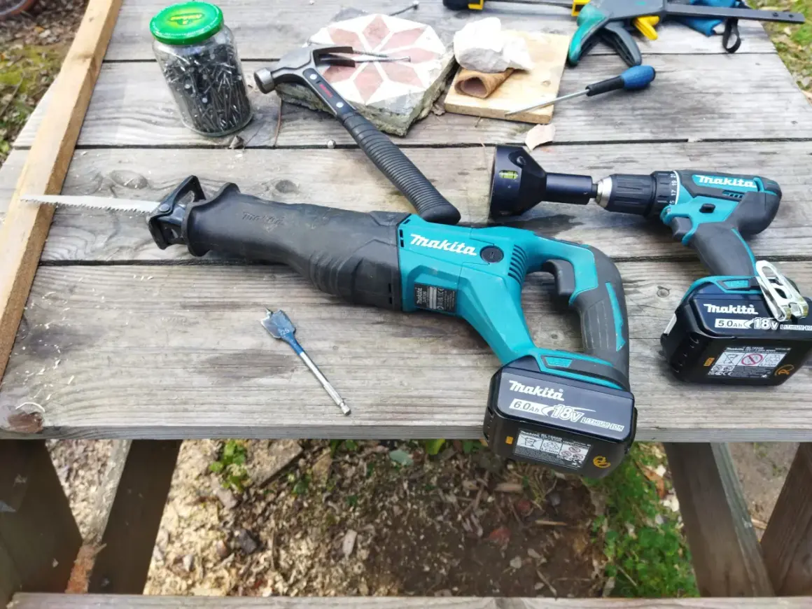 Outils Makita, marque d'outillage américain, prêts pour le travail du bois. Une scie alternative, une perceuse, un marteau et des clous sur une table en bois.