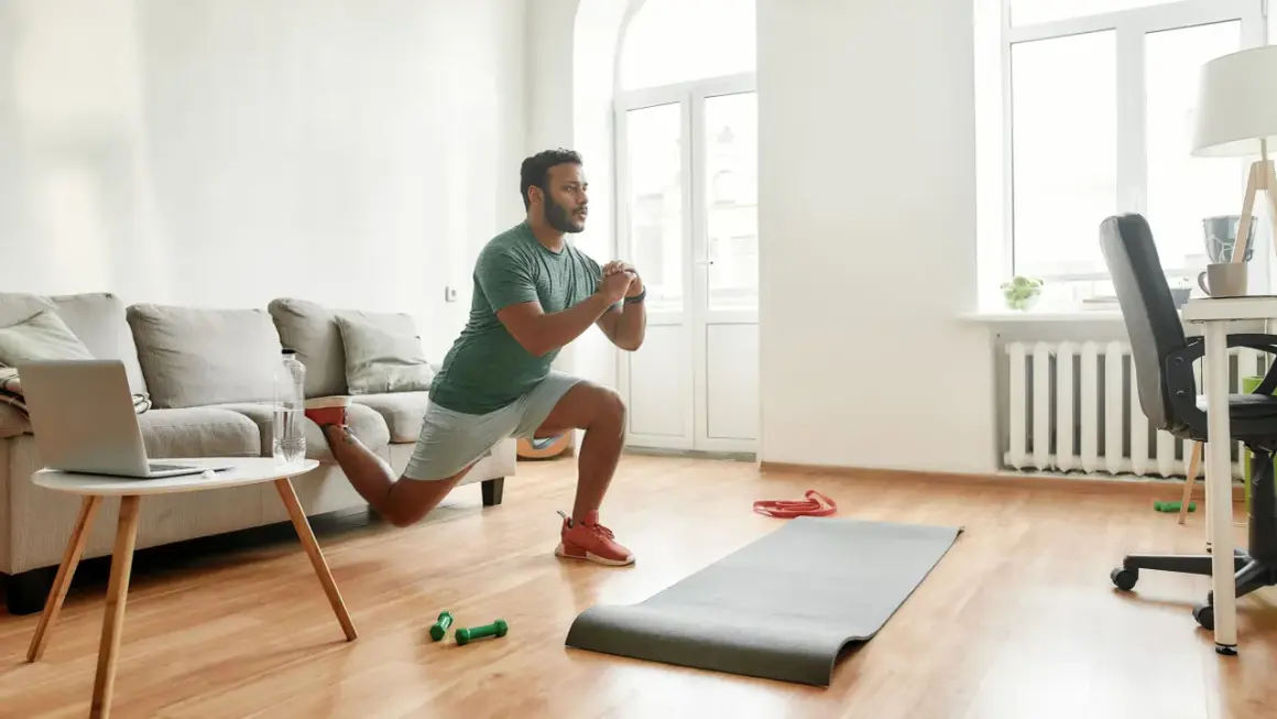 Un homme fait des fentes dans son salon, une idée pour comment commencer le sport à la maison. Un tapis, des haltères et un ordinateur sont à proximité.