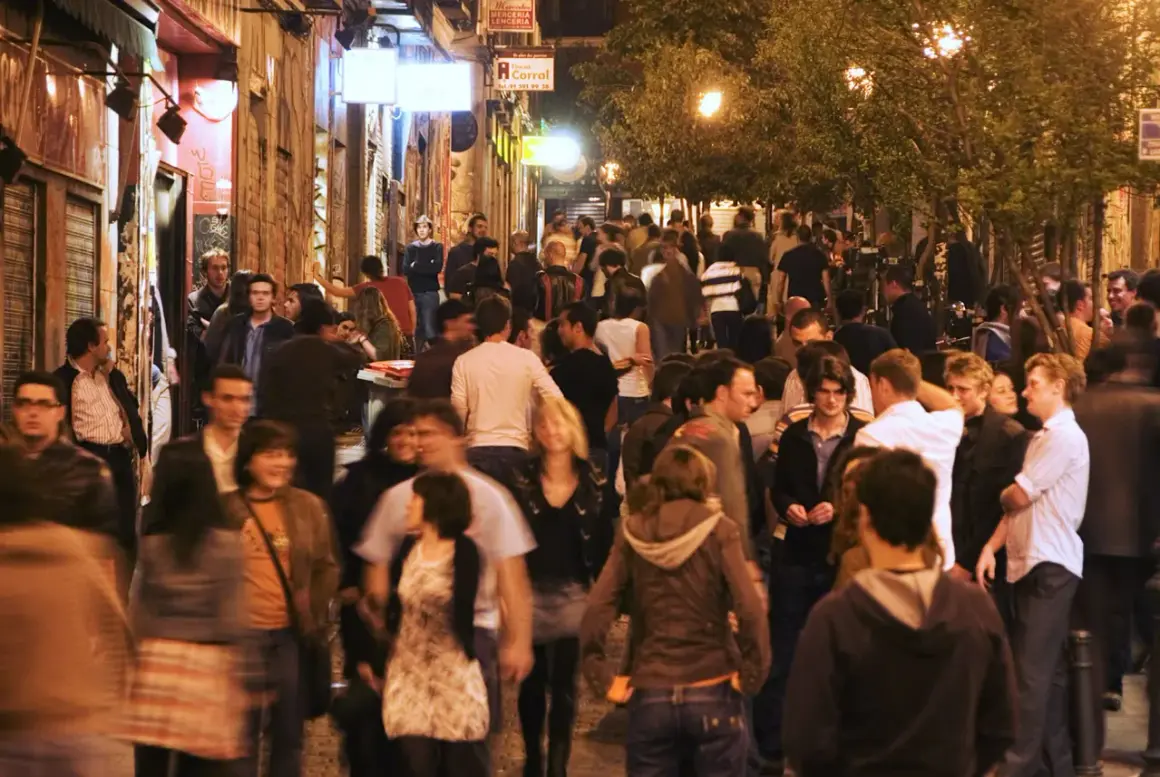 Folla animata in una stretta via di Madrid, tra luci soffuse e insegne luminose, perfetta per la movida.