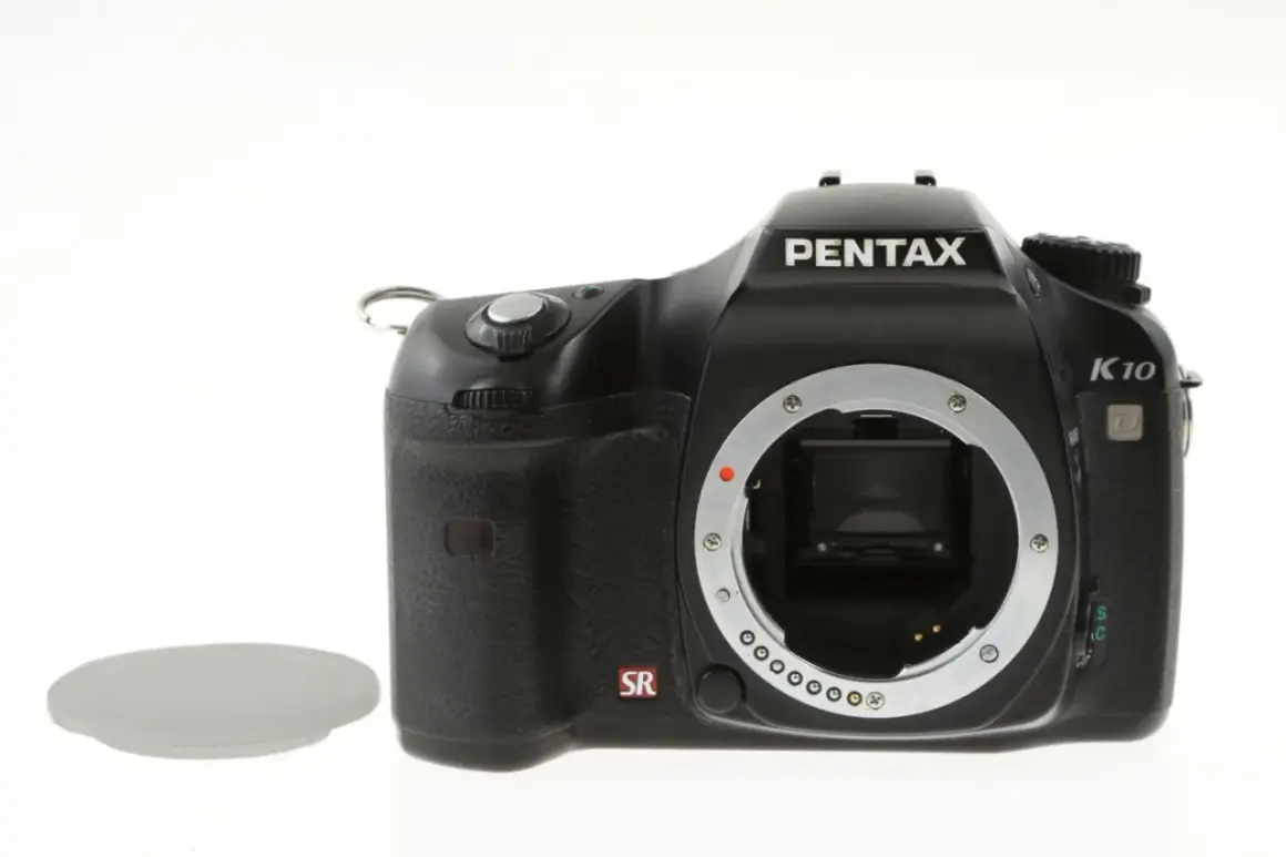 Czarny aparat Pentax K10 z widocznym mocowaniem obiektywu i symbolem SR. Obok leży biała zaślepka. Ten aparat posiada zaawansowany ccd.
