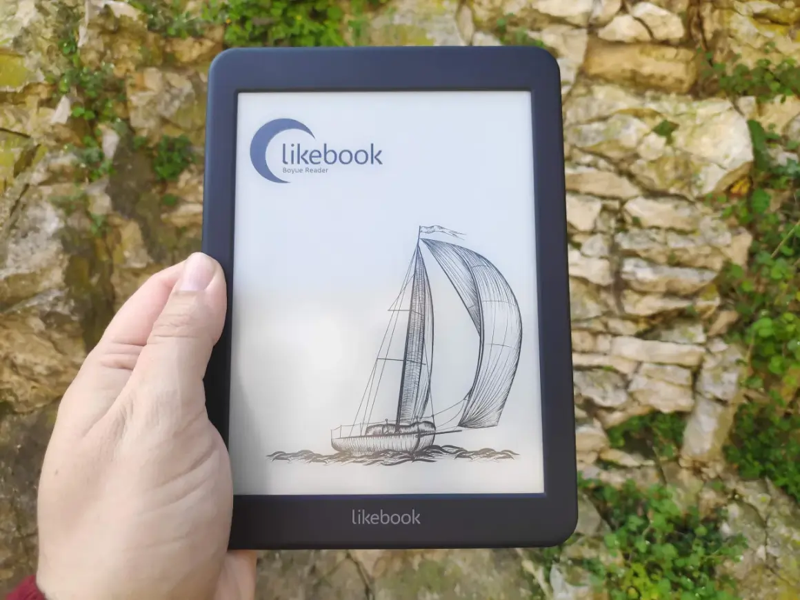 Une main tient un e-reader Likebook affichant un voilier. L'appareil est devant un mur de pierres couvert de végétation.