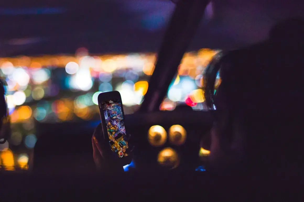 Hand hält Handy mit Stadtansicht, während sie geblitzt wird. Bokeh-Lichter im Hintergrund.