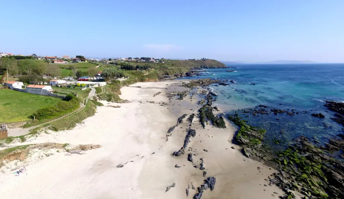 Vista aérea de la playa pragueira, con arena blanca, rocas cubiertas de algas y el mar azul turquesa. Casas y colinas verdes al fondo.