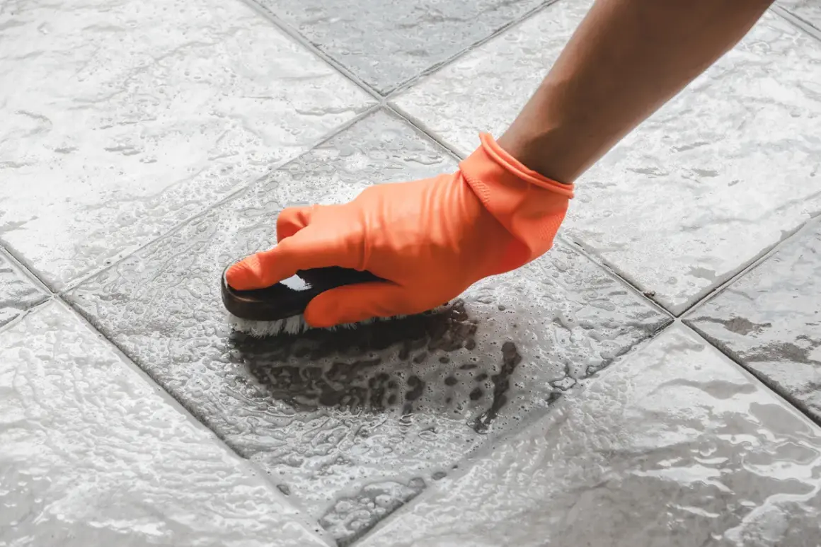 Mano con guanto arancione pulisce le fughe del pavimento con una spazzola, creando schiuma.