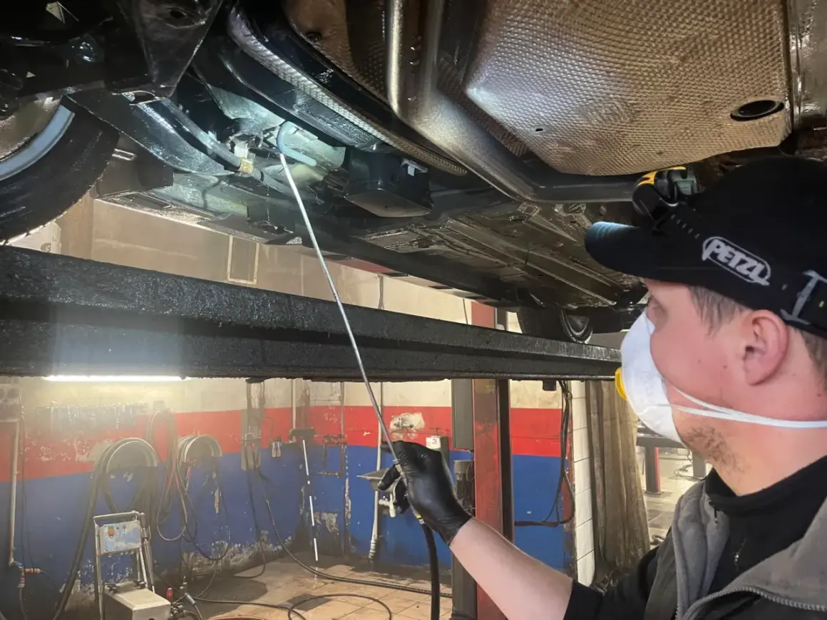 Mechanik w masce i rękawiczkach aplikuje środek konserwujący na podwozie samochodu. Długi aplikator pozwala dotrzeć do trudno dostępnych miejsc, co jest kluczowe, by wiedzieć, ile trwa konserwacja podwozia.