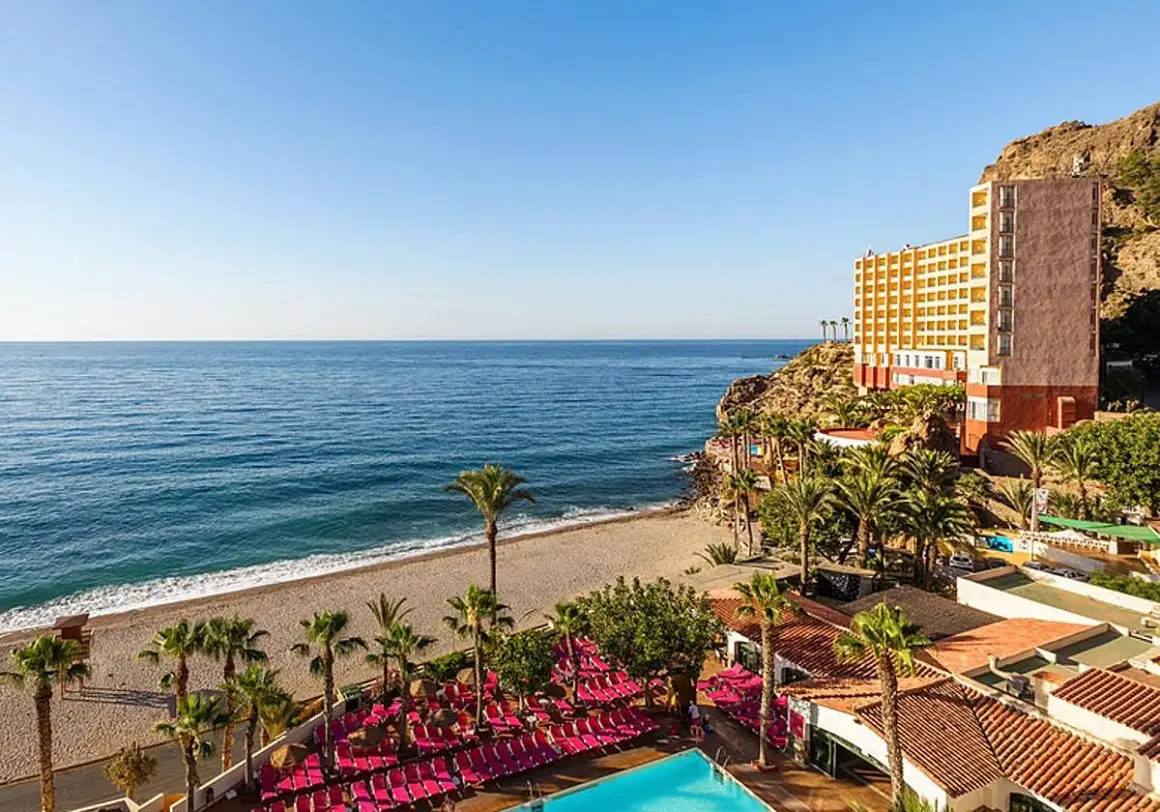 Hotel con piscina y tumbonas rosas junto a la playa de aguadulce roquetas de mar, con el mar azul de fondo.