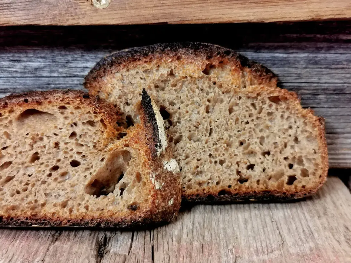 Zwei Scheiben rustikales Brot liegen auf Holz. Die Kruste ist dunkel und knusprig, das Innere luftig. Ein Beweis für gelungenes Brot backen.