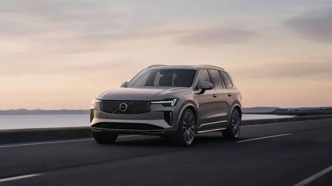Elegancki Volvo XC90 T8 w kolorze szarym jedzie wzdłuż malowniczego wybrzeża o zachodzie słońca.
