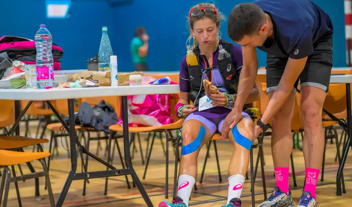 Une athlète se prépare avant une course, mangeant un sandwich. Son partenaire applique un **anti inflammatoire avant sport** sur ses genoux avec du tape bleu.