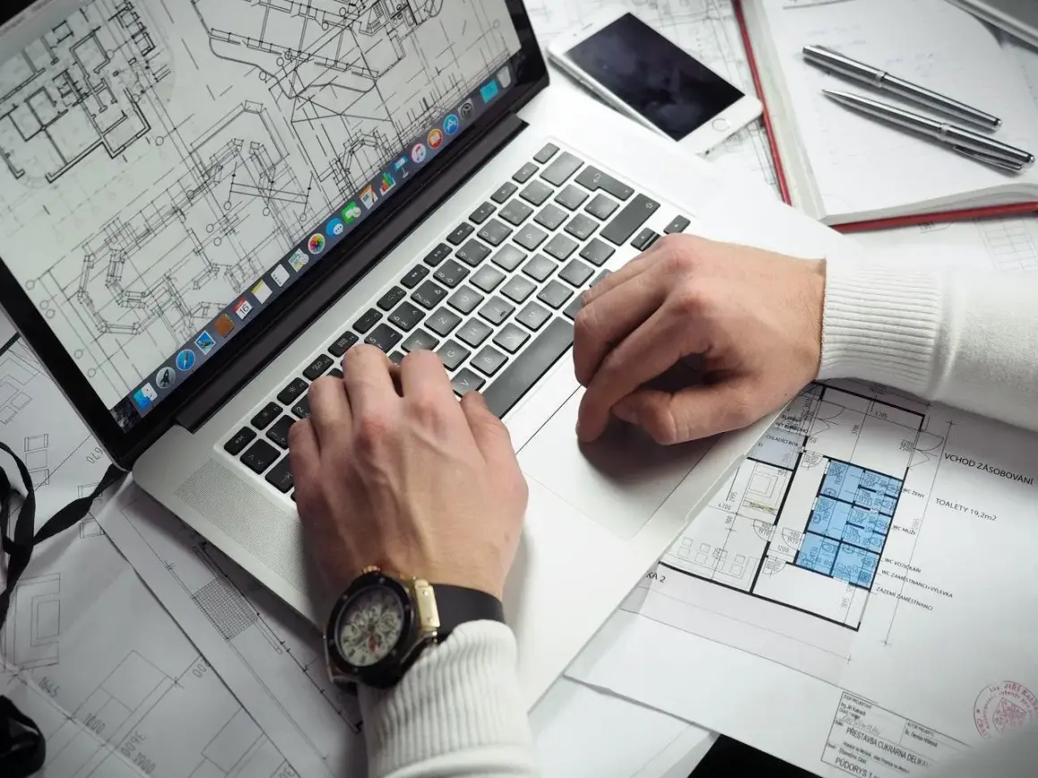 Rzeczoznawca z banku PKO analizuje plany architektoniczne na laptopie. Na biurku leży też telefon i długopisy.