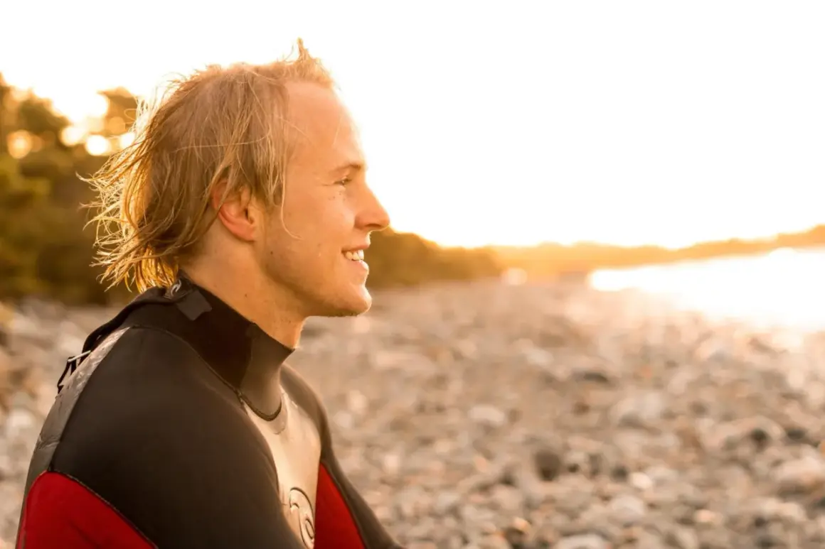 Uśmiechnięty surfer w piance, na planie średnim, patrzy w stronę zachodzącego słońca na kamienistej plaży.