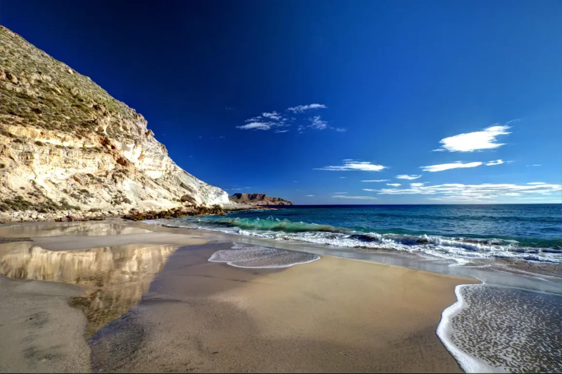 Playa de arena dorada y acantilados rocosos en la cala del plomo, con el mar azul intenso y olas espumosas.