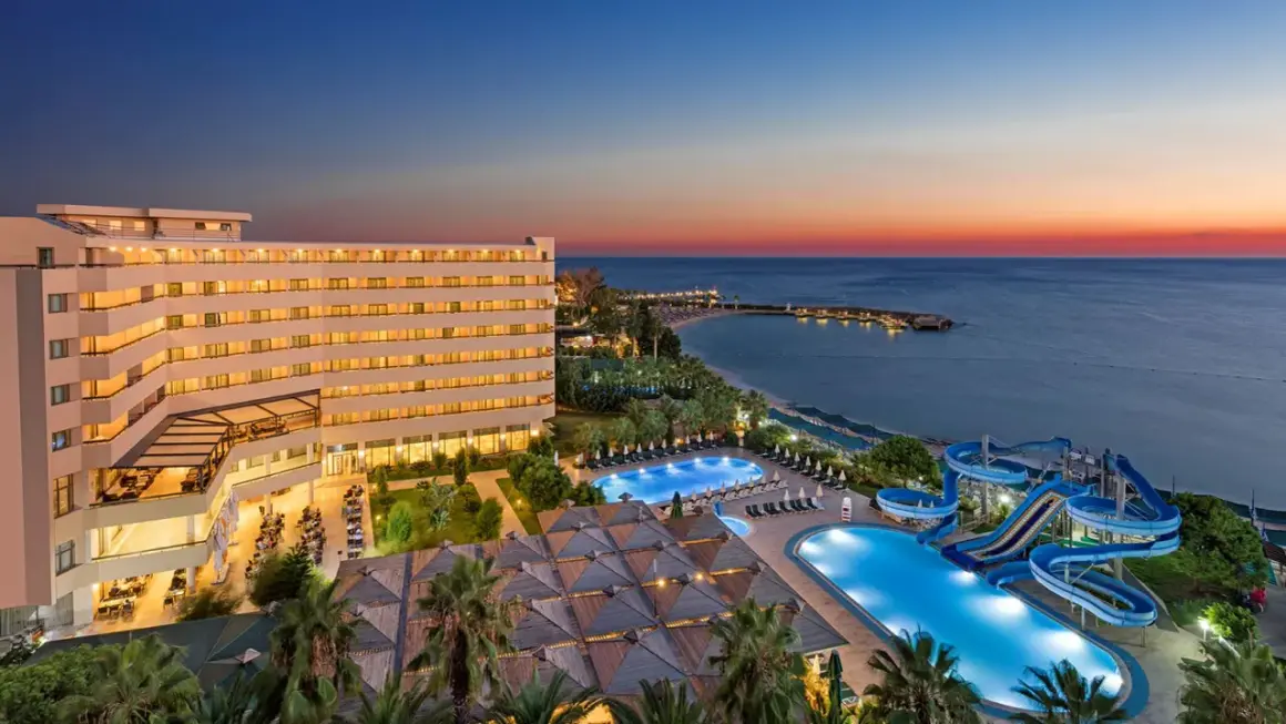 Oświetlony hotel Ozkaymak Select Resort Hotel z basenami i zjeżdżalniami wodnymi nad morzem o zachodzie słońca.
