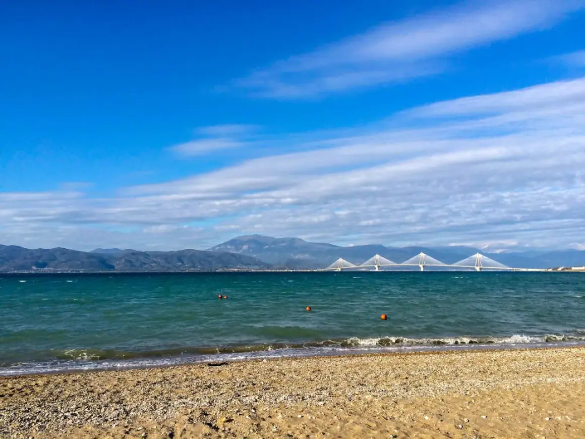 Spokojne morze i plaża w Grecji w styczniu. W oddali widać most i góry.