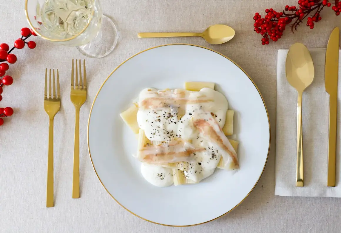 Piatto di paccheri conditi con una cremosa spuma di pecorino e guarniti con pepe nero. Accanto, posate dorate e un rametto di bacche rosse.
